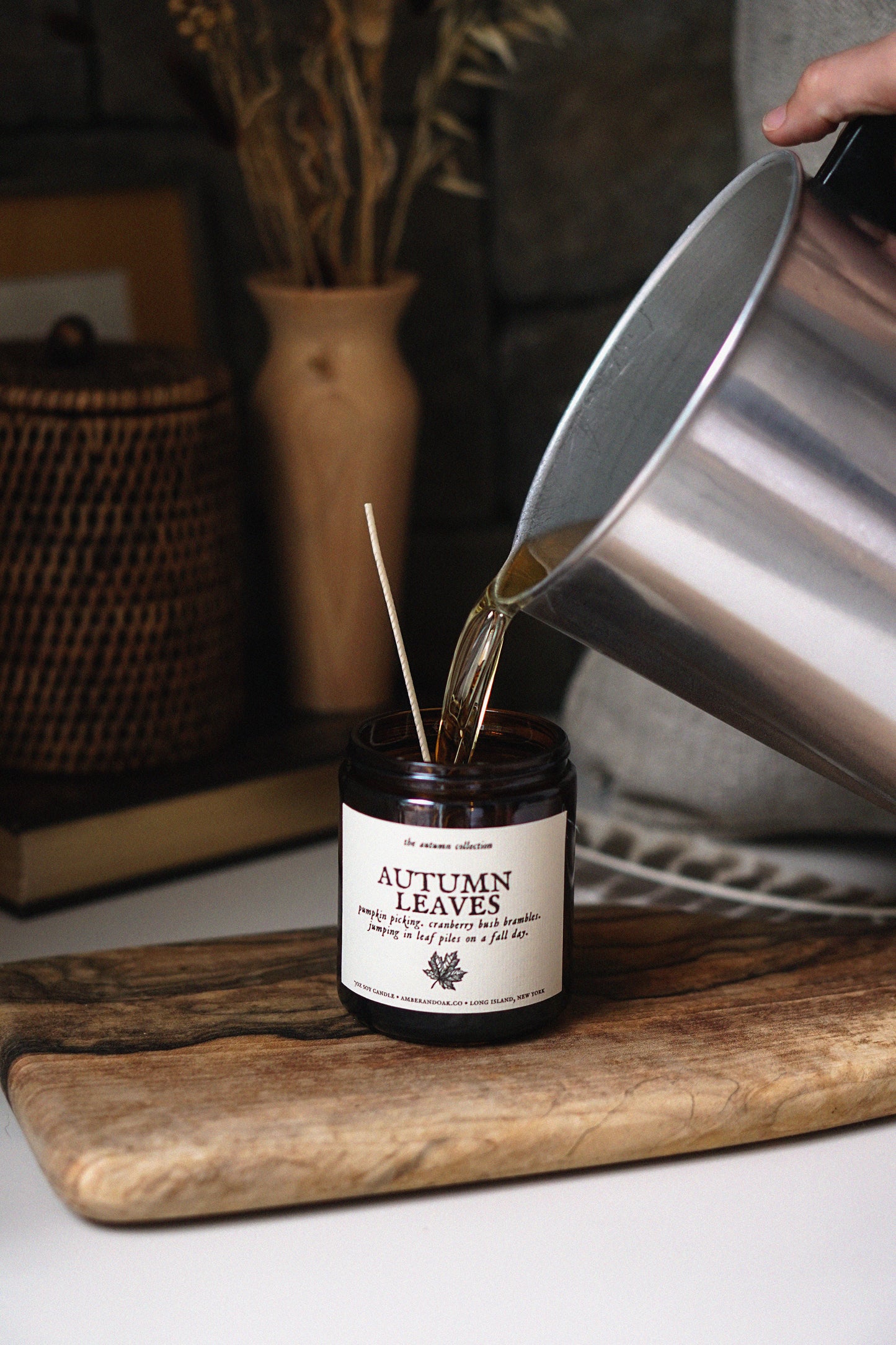 A dark amber glass jar candle labeled 'Autumn Leaves' sits on a wooden cutting board while a hand pours warm liquid from a stainless steel pot into it. The scene is styled with dried flowers, a woven basket, and a gray blanket in a cozy indoor setting.
