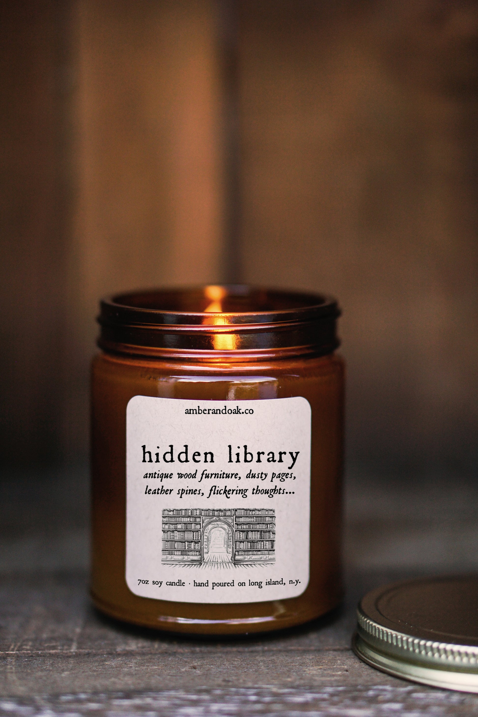 a 9 oz amber glass jar candle with soy wax and clean fragrance oils. it smells like antique wood furniture, dusty pages, leather spines, and flickering thoughts. a square kraft label fixed to the front of the jar reads "hidden library" followed by a black ink drawing of a library wall full of books. it has a gold lid and single wick.