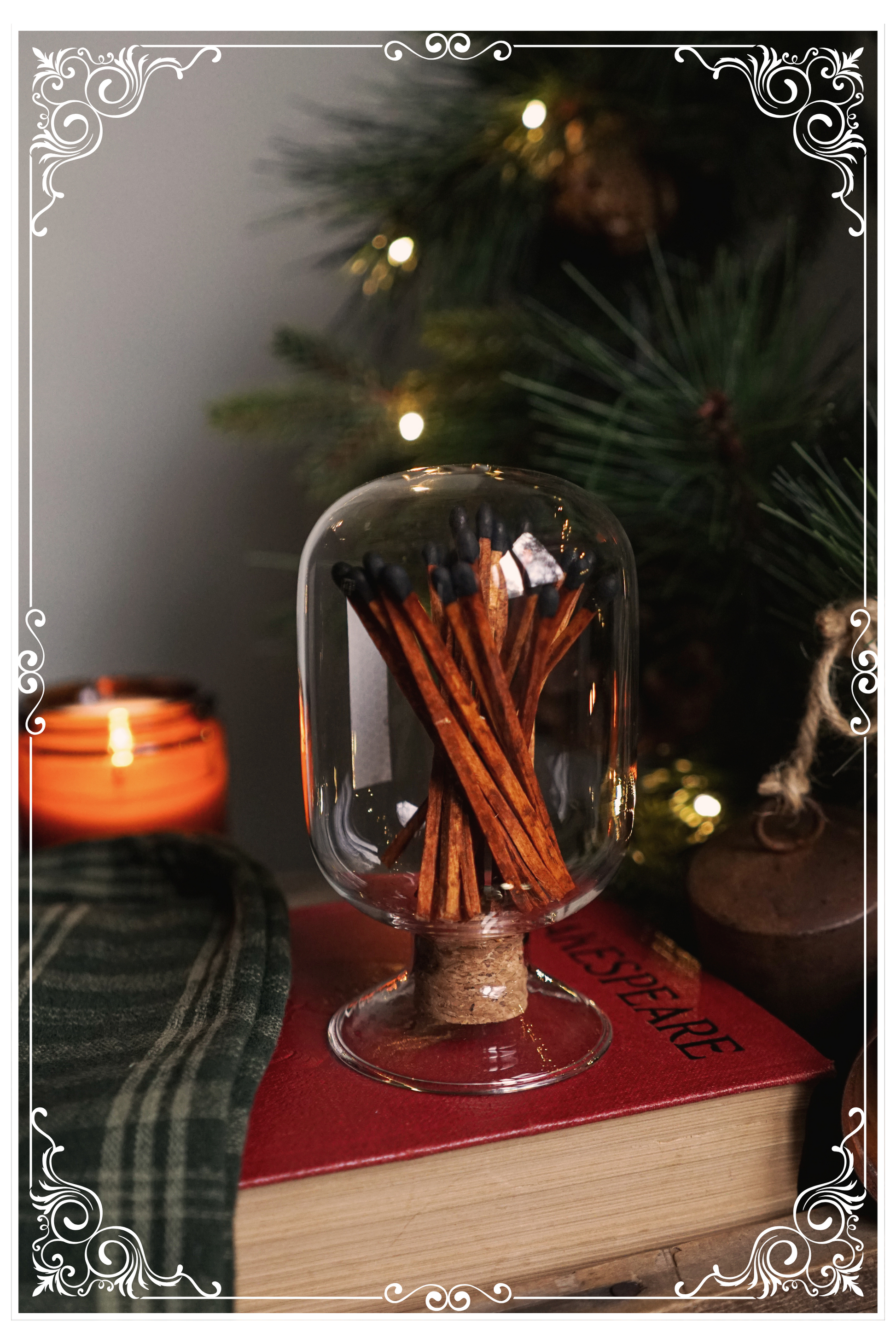 a glass cloche with dark wooden matches inside sitting atop of a book with a lit candle and evergreens in the background