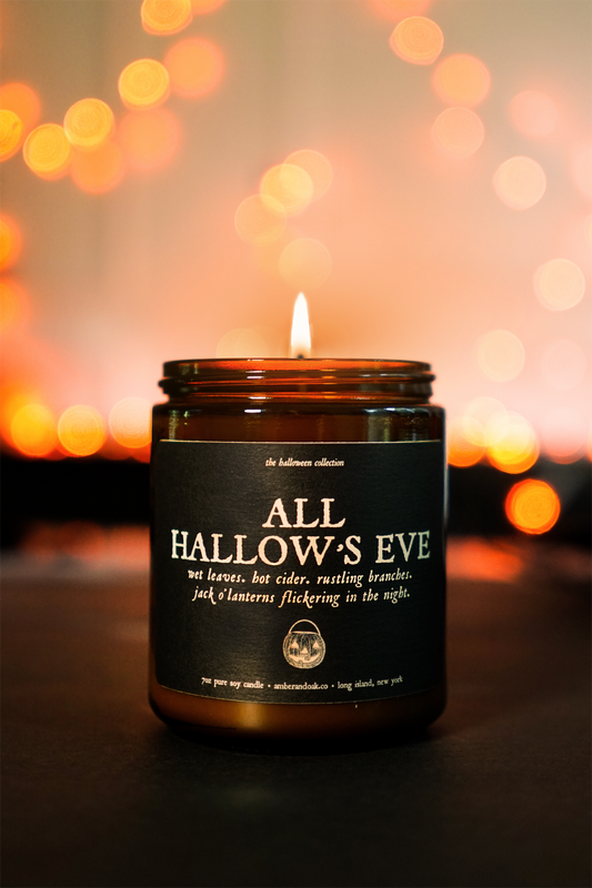 a lit amber jar candle sits in front of glowing orange lights with a black textured label that reads "all hallows eve"