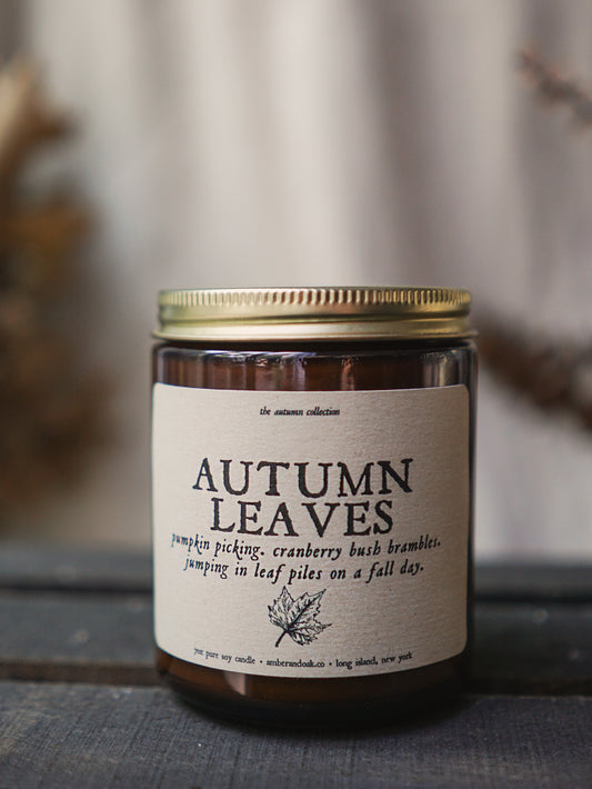 a 9 oz amber glass jar candle with soy wax and clean fragrance oils. it smells like pumpkin picking, cranberry bush brambles, and jumping in leaf piles on a fall day. a textured linen kraft colored label is fixed to the middle front with "autumn leaves" and a small drawing of a maple leaf on it.