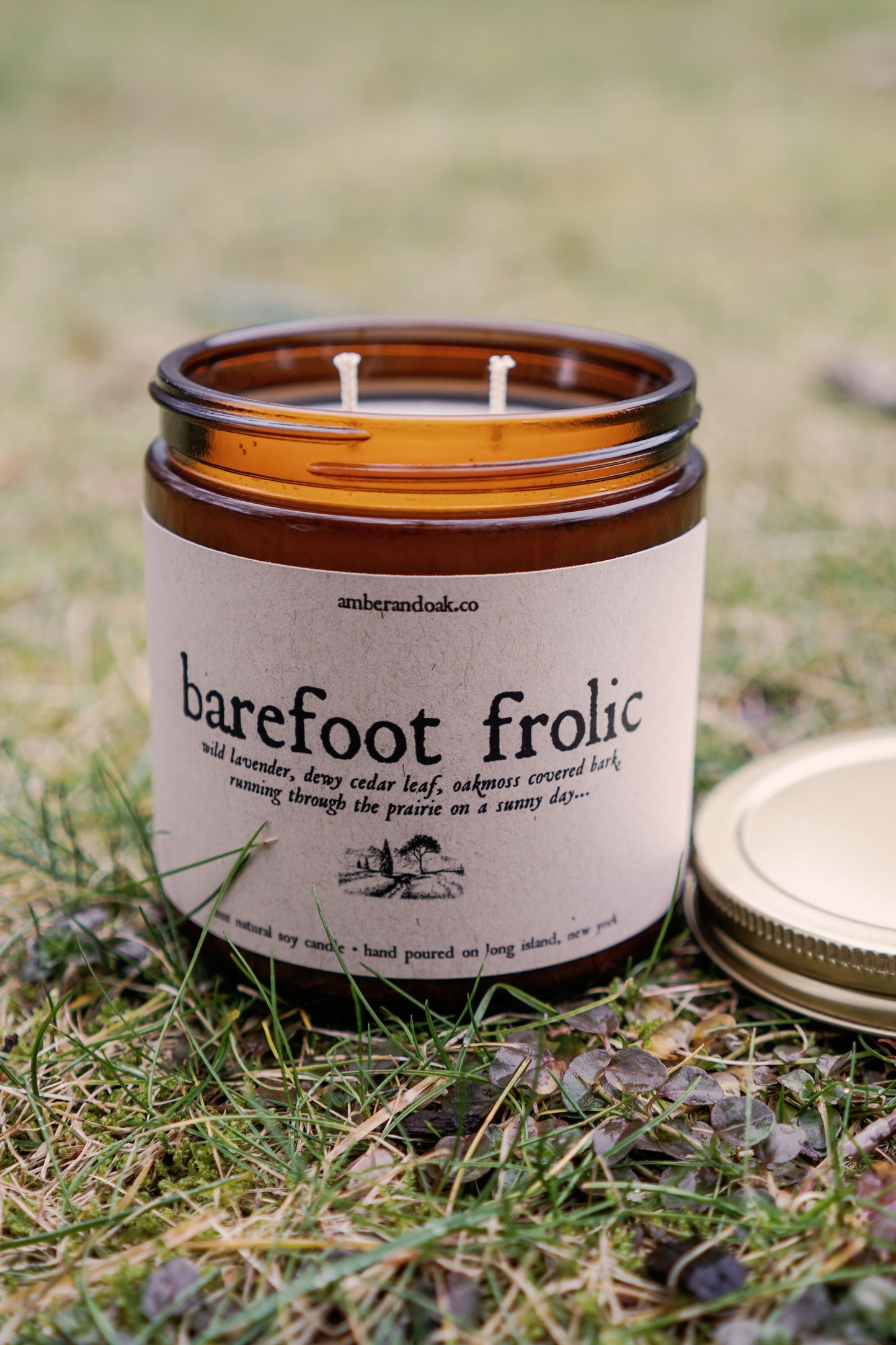 a 12 oz amber glass jar candle with soy wax and clean fragrance oils. it smells like wild lavender, cedar leaf oakmoss covered bark and running through the prairie on a sunny day. It has two wicks and a rectangular kraft label fixed to the front with the name Barefoot Frolic a drawing on a field with mountains far away in the back.