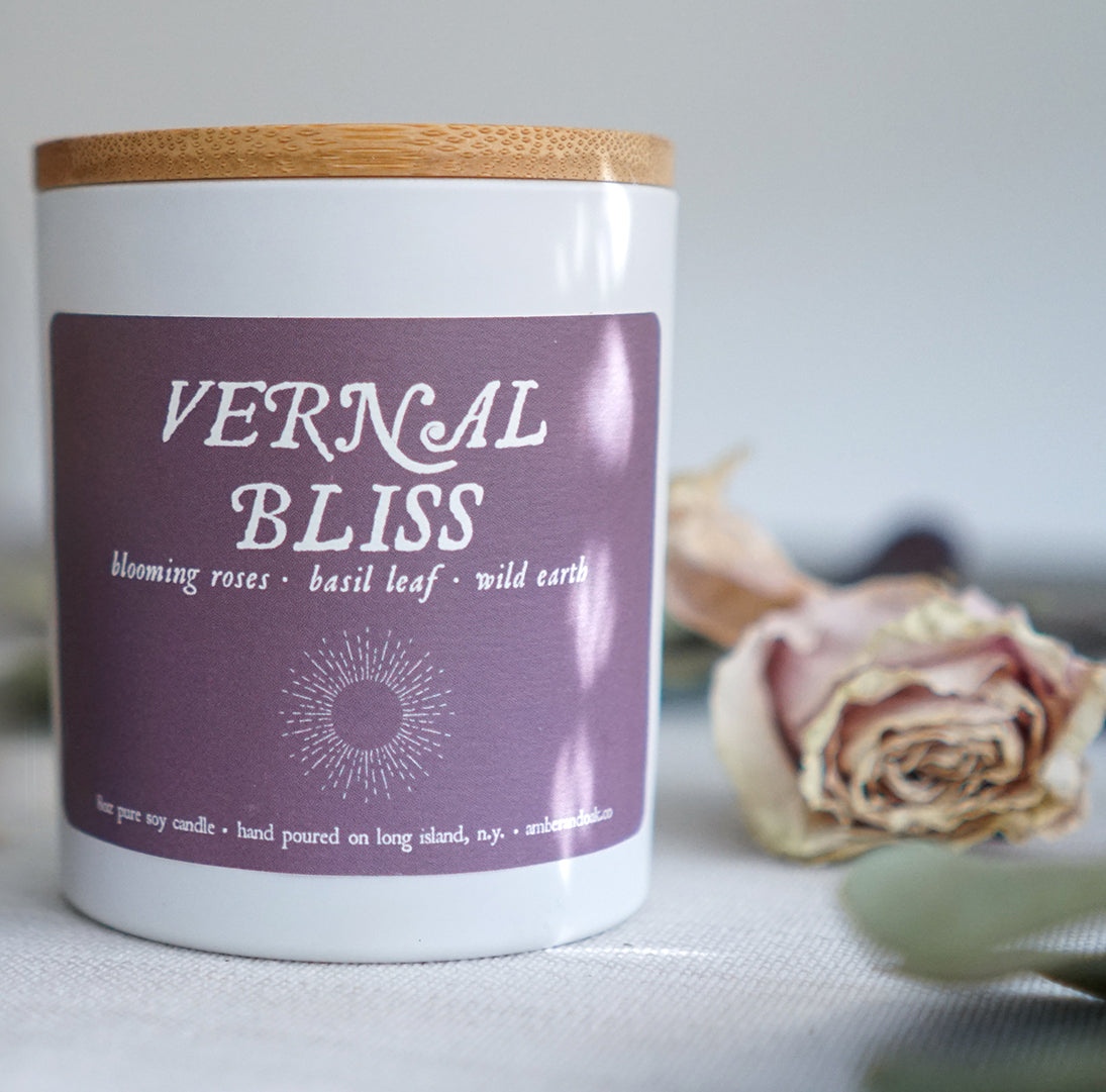 a 8 oz white ceramic jar candle with soy wax and clean fragrance oils. it smells like blooming roses, basil leaf and wild earth. a purple label with textured finish is fixed to the front reading "vernal bliss" and it has a bamboo wood lid.