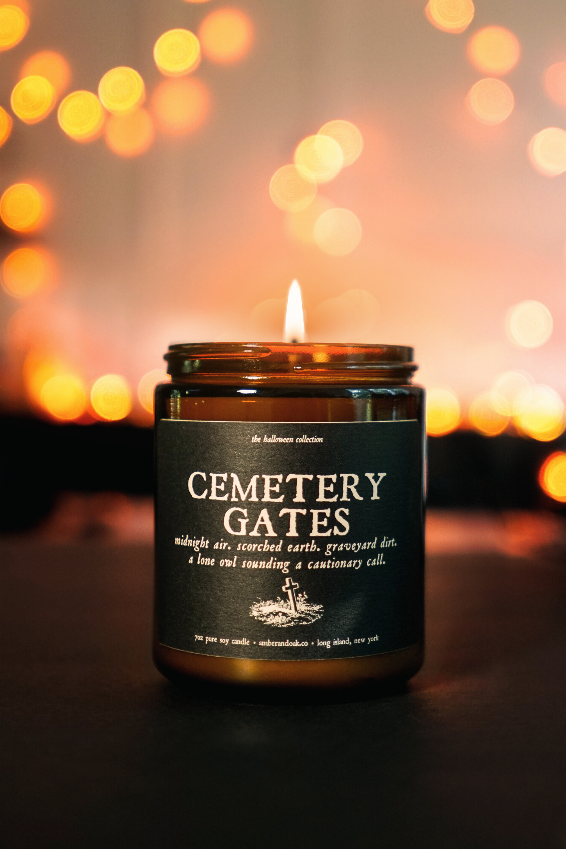 an amber jar candle sits in front of glowing orange lights. the textured black paper label reads "cemetery gates".