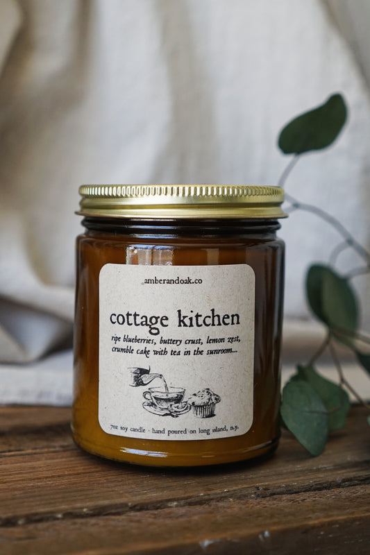 a 9 oz amber glass jar candle with soy wax and clean fragrance oils. it smells like ripe blueberries, buttery crust, lemon zest and crumble cake with tea in the sunroom. A square kraft label fixed to the front of the jar reads "cottage kitchen" with a drawing of tea being poured into a tea cup next to a muffin. It is all in black in and has a rustic primitive look to it. The candle has a gold lid.