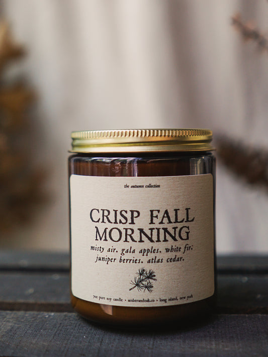 a 9 oz amber glass jar candle with soy wax and clean fragrance oils. it smells like misty air, gala apples, white fir, juniper berries, and atlas cedar. a textured linen kraft label is fixed to the front of the jar and says "crisp fall morning" followed by a black ink drawing of an evergreen branch. It has a gold lid and one wick.