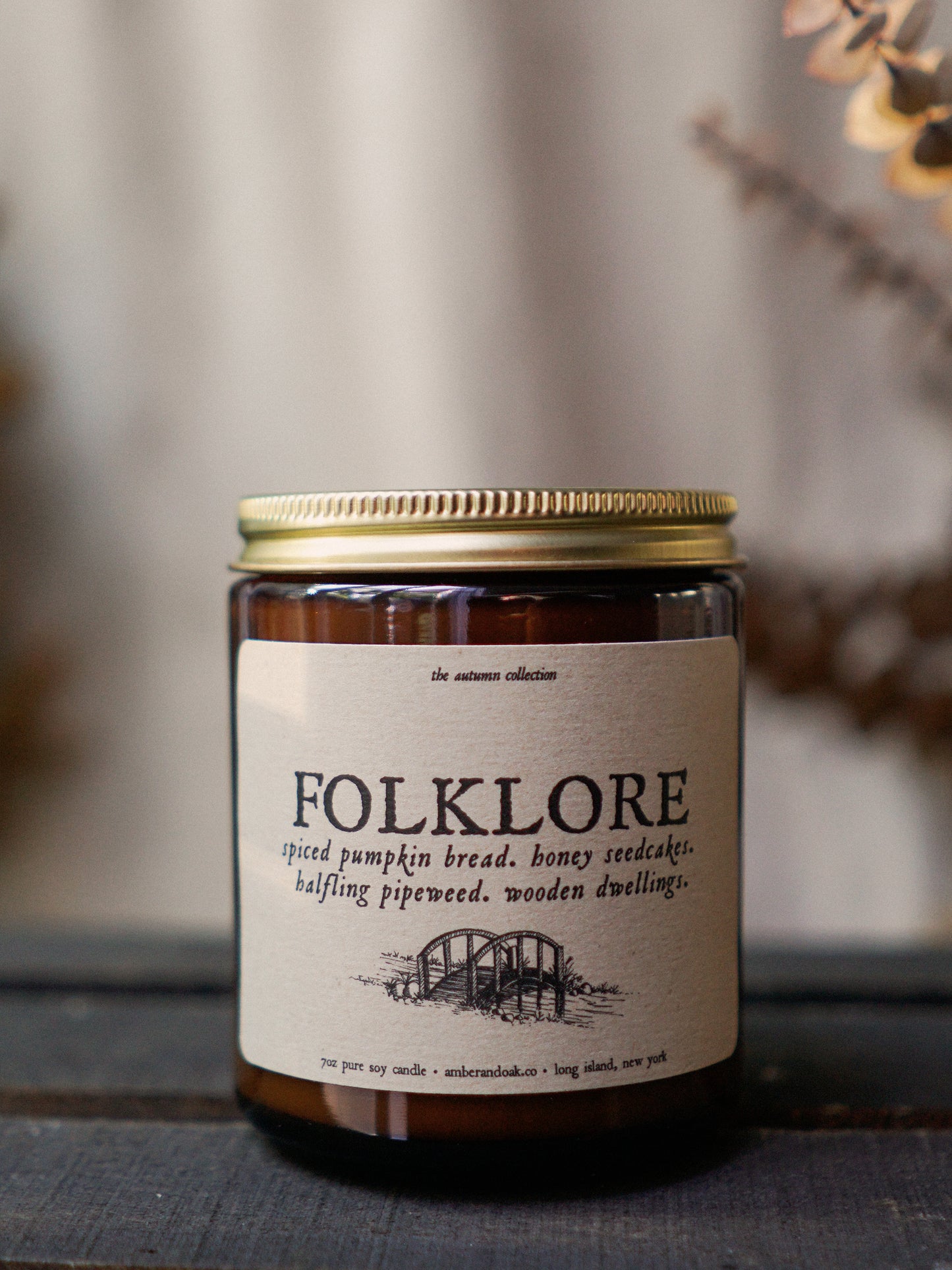 a 9 oz amber glass jar candle with soy wax and clean fragrance oils. it smells like spiced pumpkin bread, honey seedcakes, halfling pipeweed and wooden dwellings. There is a textured kraft label fixed to the front of the jar with the name "folklore" and a black ink drawing of a bridge. It has a gold lid and one wick.