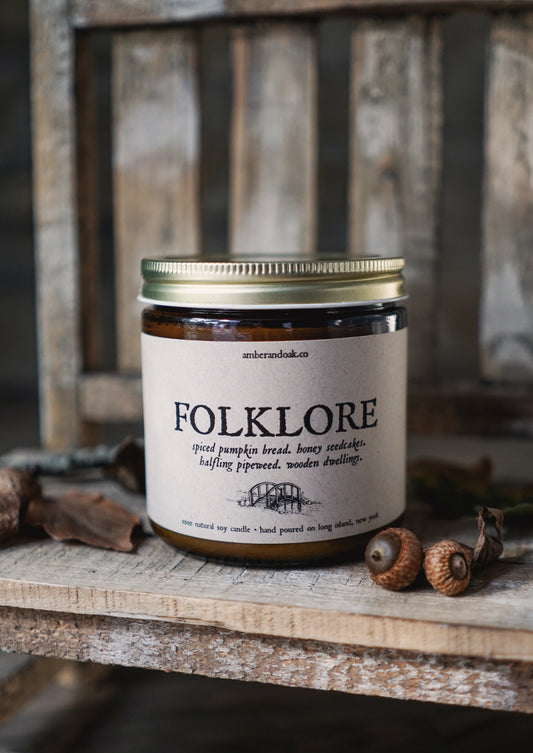 an amber jar candle sits atop of a rustic bench covered in acorns and fall leaves. The light hits it softly during golden hour and the kraft brown label reads "folklore. spiced pumpkin bread, honey seedcakes, halfling pipeweed, wooden dwellings." it has two wicks.