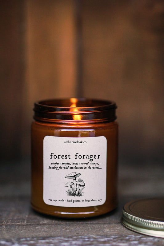 a 9 oz amber glass jar candle with soy wax and clean fragrance oils. it smells like conifer canopies, moss covered stumps, and hunting for wild mushrooms in the woods. a square kraft label is fixed to the front of the jar and says "forest forager" followed by a black ink drawing of mushrooms. It has a gold lid and one wick.