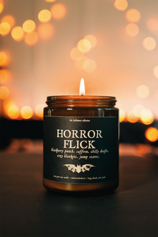 an amber jar candle sits in front of glowing orange lights. it's textured black paper label reads "horror flick"