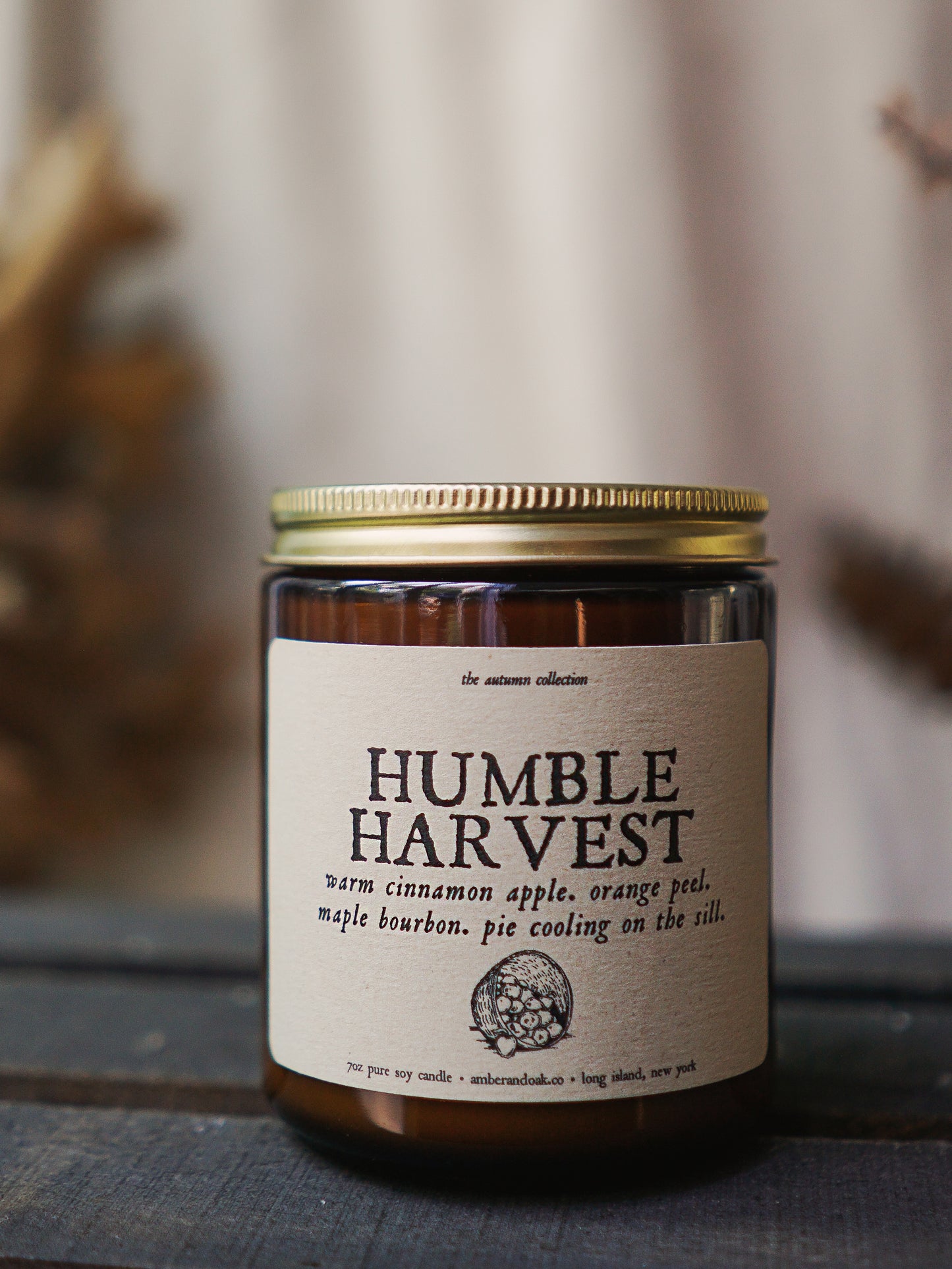 a 9 oz amber glass jar candle with soy wax and clean fragrance oils. it smells like warn cinnamon apples, orange peel, maple bourbon and pie cooling on the sill. it has a textured linen kraft label with "humble harvest" written in a rustic font followed by a picture of a basket of apples tipped over. it has a gold lid and single wick.