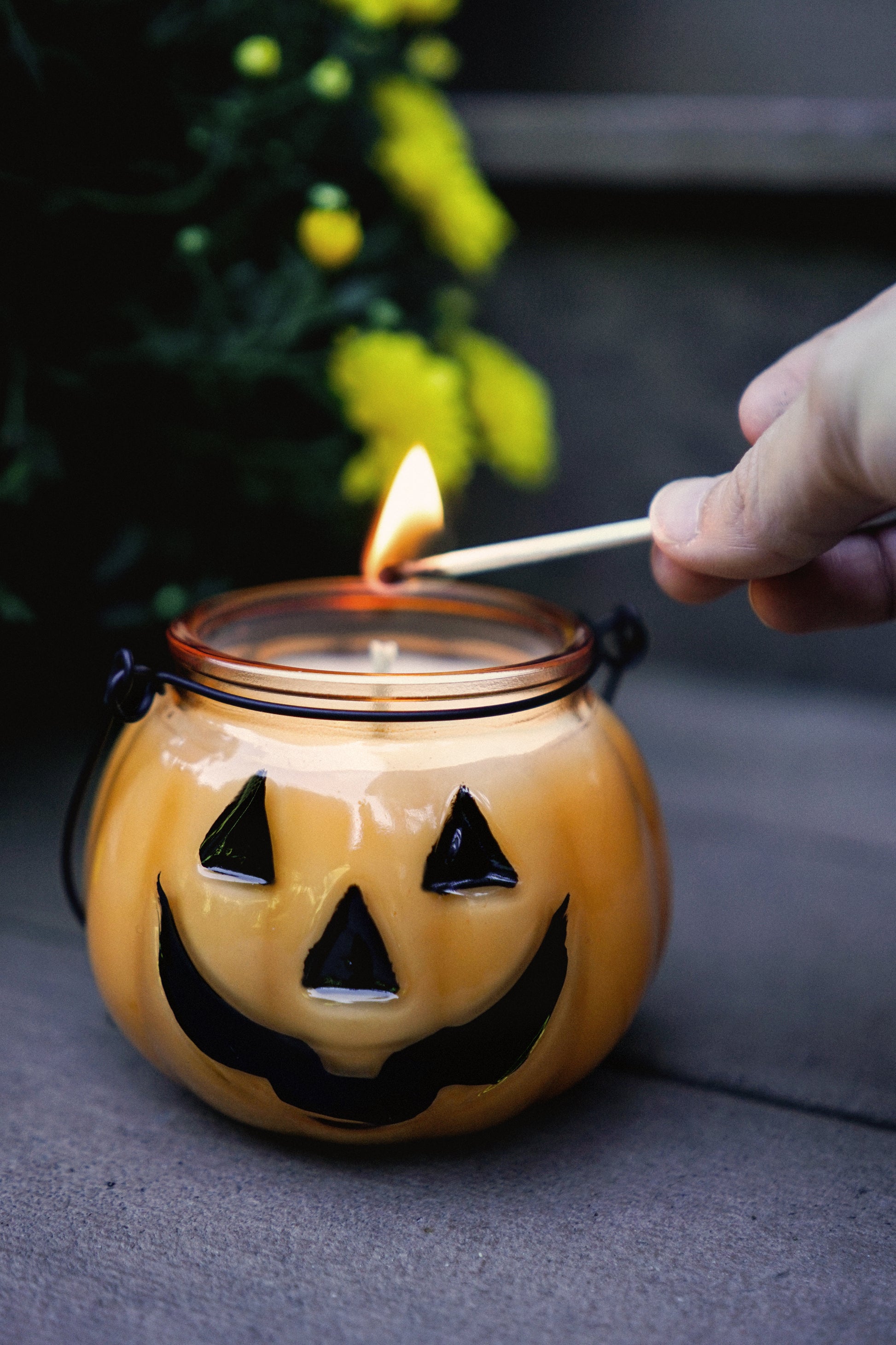 a glass orange jack o lantern candle with black triangle eyes, a triangle nose and smiling mouth. It has a black handle made of metal to carry it and smells like falling leaves + apple cider donuts in autumn. In the photo, a hand is seen with a match lighting the single wick of this pumpkin shaped natural soy candle.