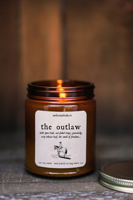 a 9 oz amber glass jar candle with a kraft square label on it saying "the outlaw, wide open land, sun faded chaps, gunsmoke, crisp tobacco leaf, the smell of freedom" in black ink followed by a drawing of a cowboy on a horse facing away. the candle is lit and a gold lid sits nearby.