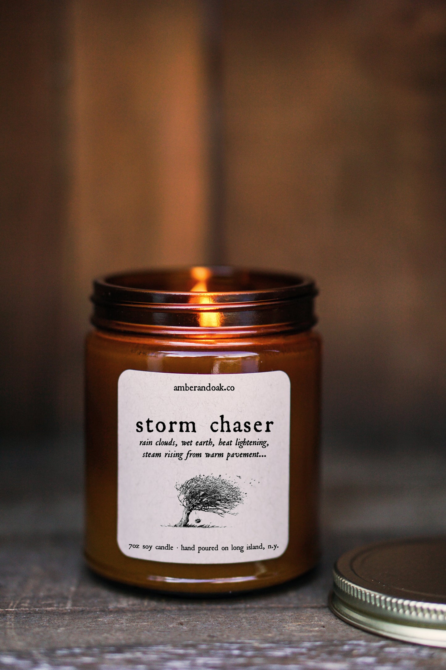 a lit candle in a 9 oz amber glass jar with a square kraft label reading "storm chaser" in black ink followed by a drawing of a tree blowing in stormy winds. 