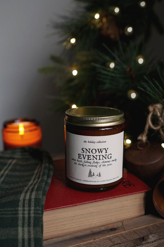 snowy evening candle sitting atop a red book with a lit evergreen and candle in the back