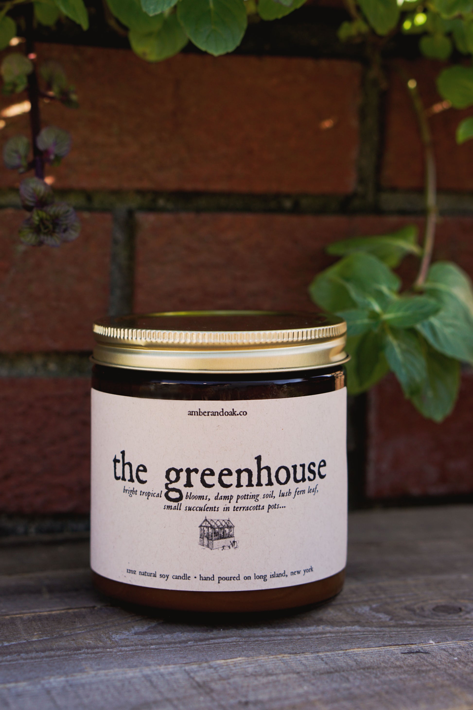 a large two wick natural soy candle in an amber glass jar with a kraft rectangular label fixed to the front reading "the greenhouse, tropical blooms, damp potting soil, fern leaf, small succulents in terracotta pots" in black ink followed by a drawing of a greenhouse. it has a gold lid. 