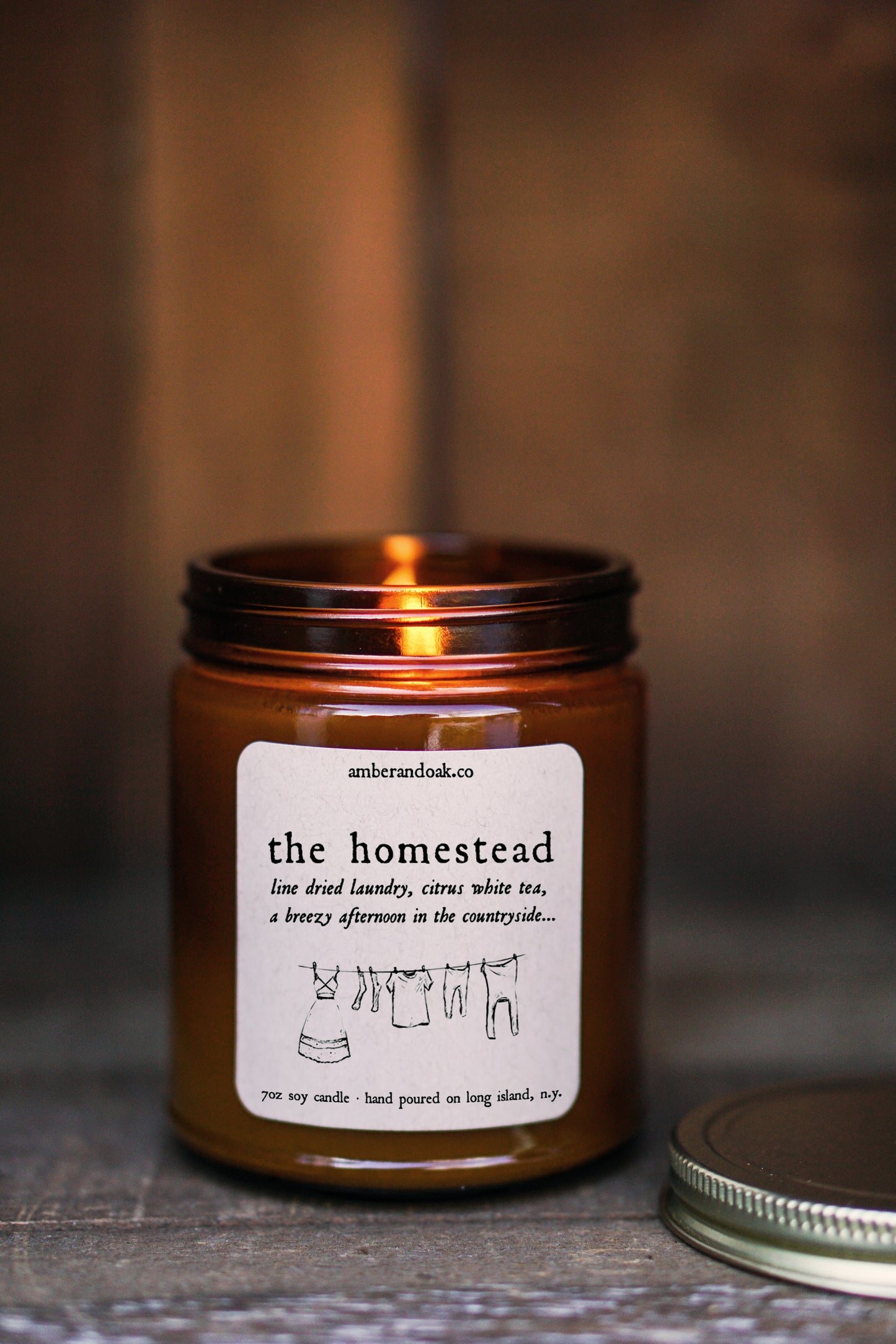 a 9oz amber glass jar candle with a kraft square label on it reading "the homestead, line dried laundry, citrus white tea, a breezy afternoon in the countryside". It has a gold lid .