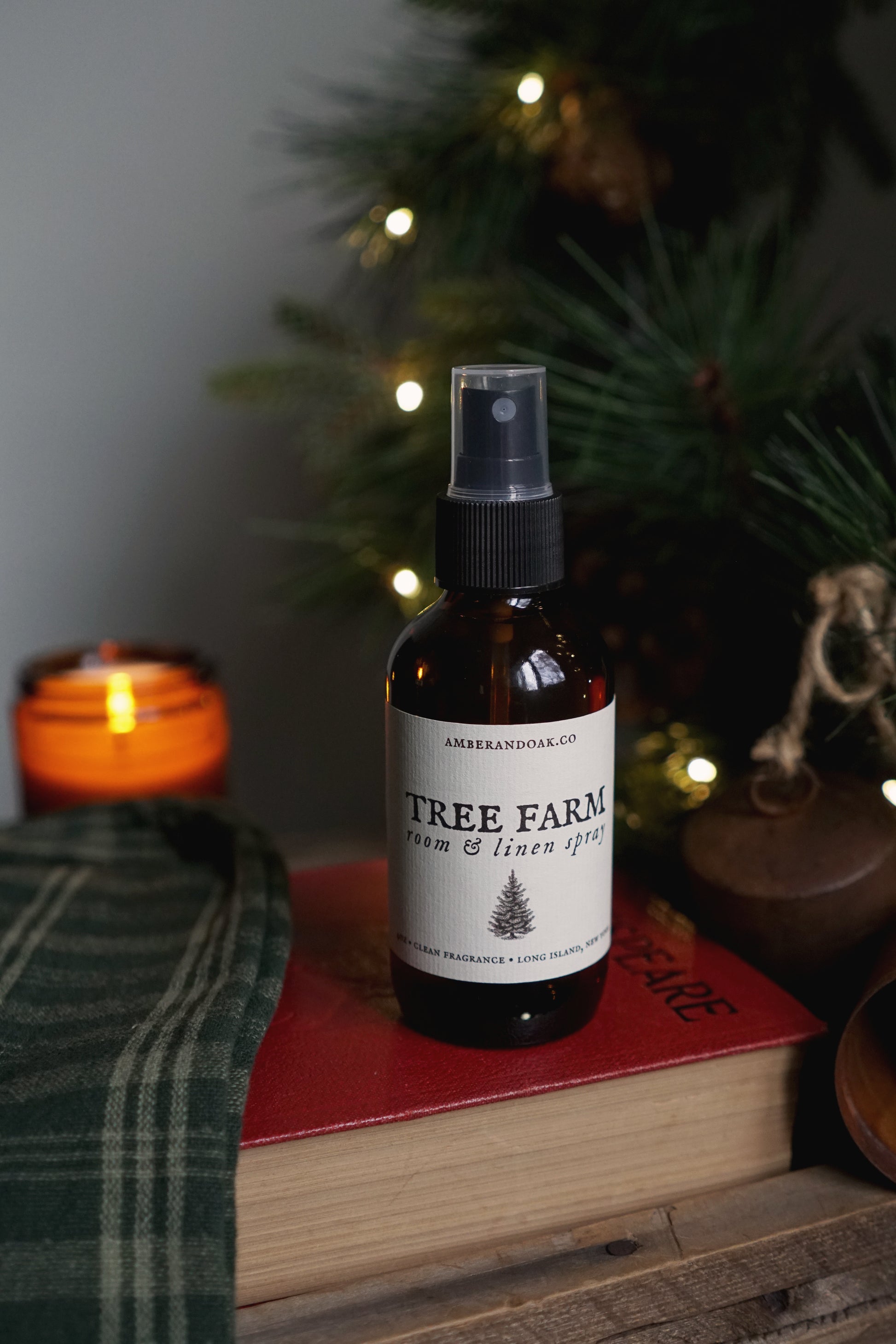 an amber glass spray bottle with a tree farm room spray label on it, sitting on top of a book with lit evergreen and candle in the background