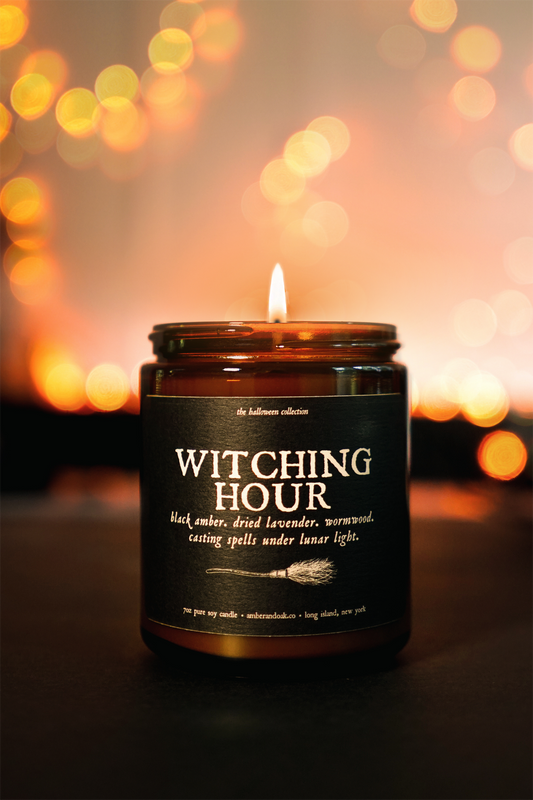 an amber jar candle with a lit wick sits in front of spooky orange glowing lights. the label is textured black paper and says "witching hour"