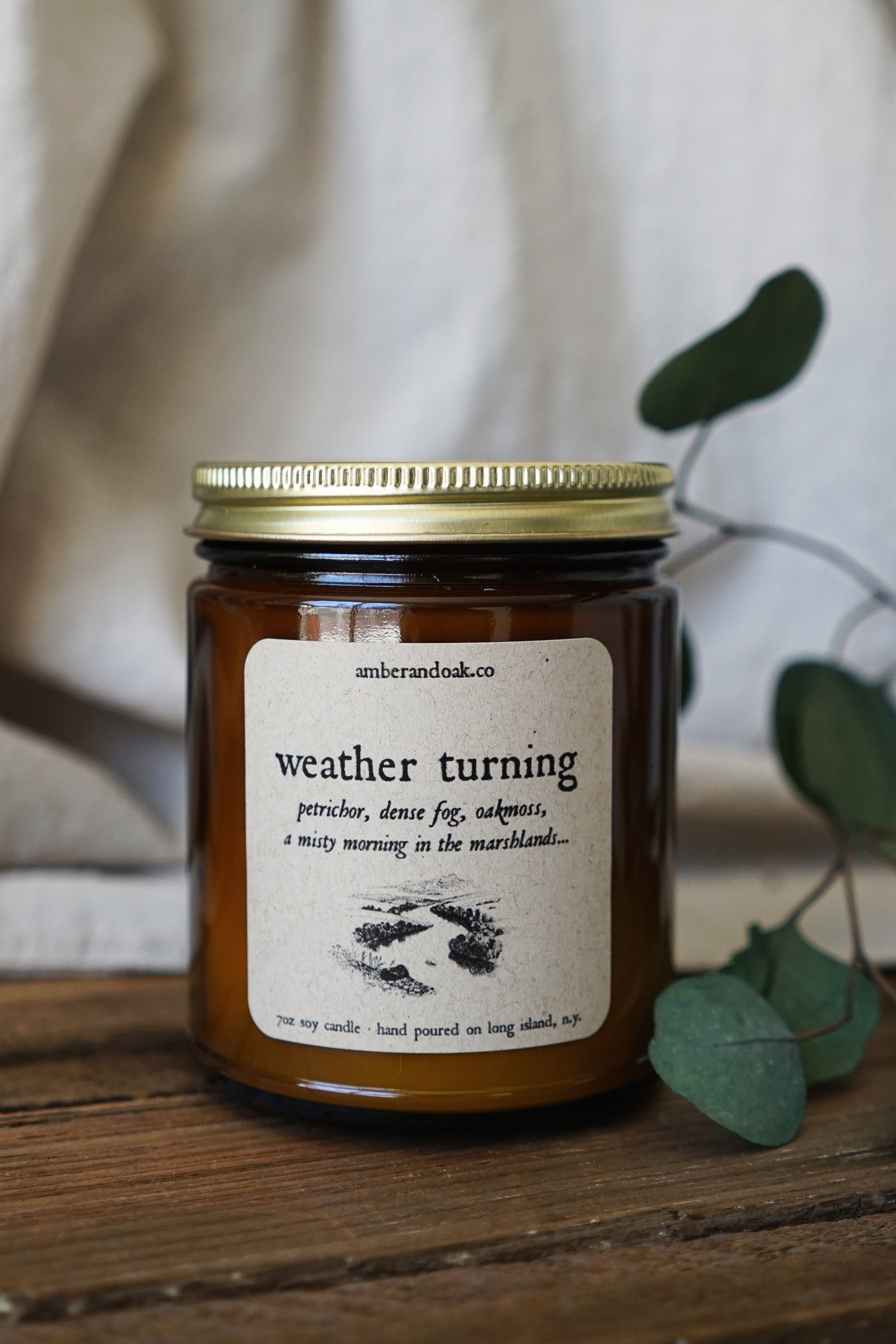 a 9oz amber glass jar candle with a square kraft label fixed to the front reading "weather turning, petrichor, dense fog, oakmoss, a misty morning in the marshlands" in black ink followed by a drawing of a river with marshland flora around it. It has a gold lid.
